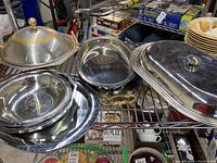 Metal serving trays and bowls arranged on a shelf, showing brass-trimmed lidded serving bowl and various round and oval trays with handles and hammered details.