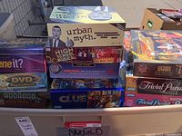 Stack of various boxed board games including Urban Myth, High School Musical DVD board game, Clue electronic game, and box edges of Trivial Pursuit.