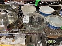 Wide view of three Pyrex bowls with lids, two blue and white Corning Ware dishes and one chipped Corning Ware dish on metal shelf with three stainless steel Tramontina steamers.