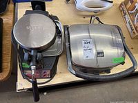 Top and angled view of the Waring waffle maker and Gordon Ramsay panini grill placed side by side on a wooden surface, showing metal finishes and handles.