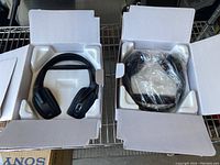 Two pairs of black wireless stereo headphones in open white boxes with foam inserts. One pair is wrapped in plastic.