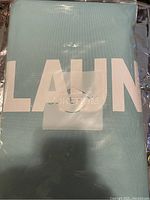 Photo of the light blue laundry bag visible inside unopened clear plastic packaging showing part of white lettering 'LAUN'.