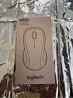Front view of the Logitech M510 Comfort Plus mouse packaging showing minimalist black line art of the mouse on brown cardboard box with Logitech logo.