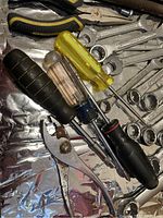 Close-up image showing 4 screwdrivers with colored handles (black, yellow, translucent), rusty adjustable pliers, and open-end wrenches beneath them.