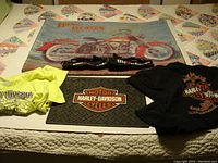 Two Harley Davidson floor mats and two Harley Davidson t-shirts displayed on a bed. Rubber mat shows standard Harley Davidson logo on textured black surface measuring 16x14 in. Carpet mat has a classic motorcycle graphic measuring 36x26 in. T-shirts are 3XL sized, one black with Harley Davidson text, the other yellow with Harley Davidson logo.