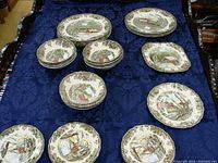 All pieces arranged on table showing dinner plates, small plates, and bowls with village scene and leaf border.