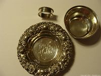 Top-down view of all three items: ornate round dish with floral border and monogram, Birks dish, and napkin ring.