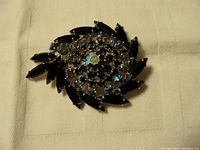 Top view of black and dark blue iridescent rhinestone vintage brooch on white cloth showing the circular swirl design.