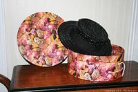 Black braided straw ladies hat with ribbon band placed beside a round floral-patterned hat box on a wooden surface.