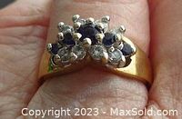 Close-up photo of the ring on a finger showing the front top view of the blue and clear gemstones set in a gilded band.