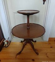 Photo showing front angled view of vintage two tier mahogany side table with pie crust edges and three legs on wooden floor.