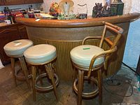 Full view of rattan bar stand with bamboo front and three cushioned bar stools, one stool with back and armrests.