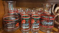 Two Coca Cola glass carafes with several matching holiday-themed Coca Cola glasses on a wooden shelf.