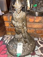 Front view of bronze-tone cowboy statue standing with a saddle, showing detailed clothing and accessories.