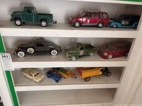 Collection of various die cast toy vehicles displayed on white shelves, includes mini vans, coupes, trucks in multiple colors and styles