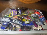 Plastic bin filled with various loose die cast toy cars and trucks including emergency vehicles and utility trucks, with assorted colors visible including blue, white, red, and yellow