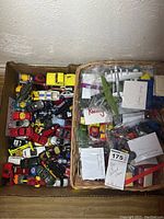 Top-down view of a basket and cardboard box filled with various die cast toy vehicles including cars, trucks, and planes.
