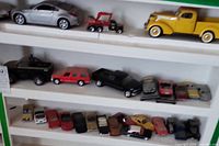 Shelf with die cast cars including silver sports car, yellow vintage pickup truck on top, multiple cars and trucks on mid and bottom shelves