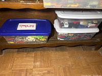 Plastic storage totes on wooden shelf labeled 'Hot Wheels' containing numerous small diecast vehicles including cars and trucks visible through translucent containers