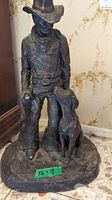 Full front view of a dark bronze-like sculpture showing man in cowboy attire with dog at his side, on a round base.