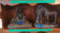 Two bronze-tone Largo sculptures placed on a wooden shelf; cowboy in a tub on the left and cowboy with horse on the right.