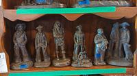 Six bronze cowboy sculptures displayed on wooden shelf, each with detailed western attire, various heights between 3 and 10 inches.