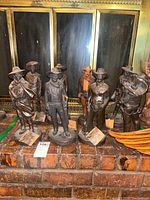 Photo of all eight bronze cowboy statues arranged on a brick shelf in front of a fireplace screen, showing various poses and detailed western attire