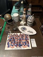 Full set of Dallas Cowboys memorabilia on table including helmets, glasses, plate, tin, cheerleader figurine, Dr. Pepper bottle, and autographed cheerleaders photo.