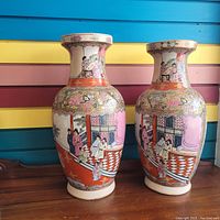 Photo showing front and left side view of two large ceramic vases with hand-painted Oriental scenes and floral motifs on a wood surface against a colorful wall.