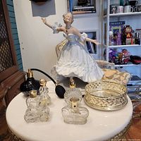 Photo shows the porcelain figurine of a woman in a white dress beside six Avon glass perfume bottles arranged on a table with a silver plated holder.