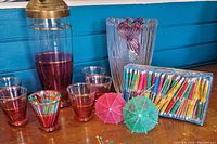Pink and gold glass cocktail shaker, six matching tinted glasses, pink and clear decorative vase, box of cocktail umbrellas and assorted drink picks laid out on table