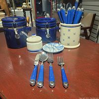 Overview of entire set showing two blue airtight canisters, open ceramic canister with blue star decorations containing blue-handled cutlery, covered jar and sailboat plate on wood table.