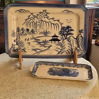 Large blue and white Bombay tray with Asian coastal scene and wooden stand along with smaller blue and white English tray with floral border and scenic center on table.