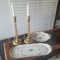 Pair of brass spiral candle holders with white candles, round serving dish and rectangular tray with gold trim on wood table