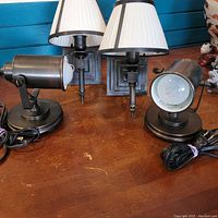 Four bronze finish lighting pieces arranged on wood surface in front of blue wall: two wall sconces with white pleated shades, two adjustable table lamps with cylindrical heads and cords coiled beside bases.