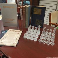 Photo of seven chess books standing upright, a 32-piece glass chess set arranged on a table, and a small booklet and star sticker sheets laid flat.