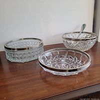 Photo showing three crystal serving pieces with silver rim, and one silver serving spoon on wooden surface, lighting highlighting cut patterns of crystal.