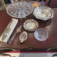 View of all seven serving pieces including glass cake stand, sterling silver bowls and dishes, glass bowl, and serving utensils