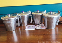Four stainless steel Haagen-Dazs ice cream holders with hinged lids placed on a wooden surface, front view with informational cards visible.
