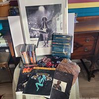 All items grouped: framed Hendrix photo, song books, hardcover coffee table book on musical instruments, t-shirts partly visible, and DVDs box sets on a chair