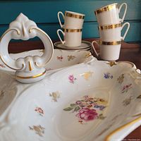 Close-up of porcelain candy dish with floral pattern and gold trim alongside stacked espresso cups with gold bands.