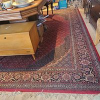 Full view of large rectangular red rug with intricate border pattern placed under furniture showing general condition and fringe