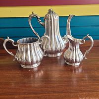Front view of three-piece silver tea set including teapot, sugar bowl, and creamer showing ribbed design and handles.
