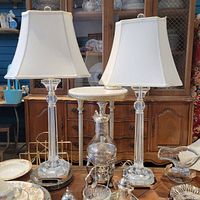Full view of two clear lucite and silver table lamps with white shades on display with other items
