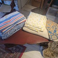 Folded bedding items on a wooden table including patterned blue and white bedding, yellow floral bedding, and a brown and blue pillowcase