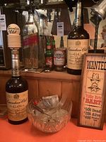 Photo shows several collector bottles including Canadian Club large bottle and Hatuey beer bottle, Budweiser can, and a wooden 'Wanted' plaque with cowboy image, plus a glass punchbowl with ladle and stein mug.