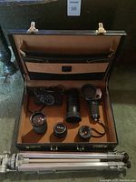 Open briefcase showing the Canon FTb camera with several lenses, Minolta AutoSpot light meter, and aluminum tripod placed below the case.