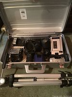 Metal case open showing multiple cameras including Pentax SF1, Hanimax, Minolta Auto Spot, Canon Elura camcorder, Sure Shot Prima, tripod, and accessories.