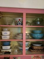 Overall cabinet view showing clear glass bowls, tulip-pattern bowl, CorningWare dishes, Pyrex bowls and wicker trays