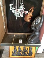 Portrait painting of a woman with a large hibiscus flower in hair, framed in wood, next to dark gray figurative sculpture.